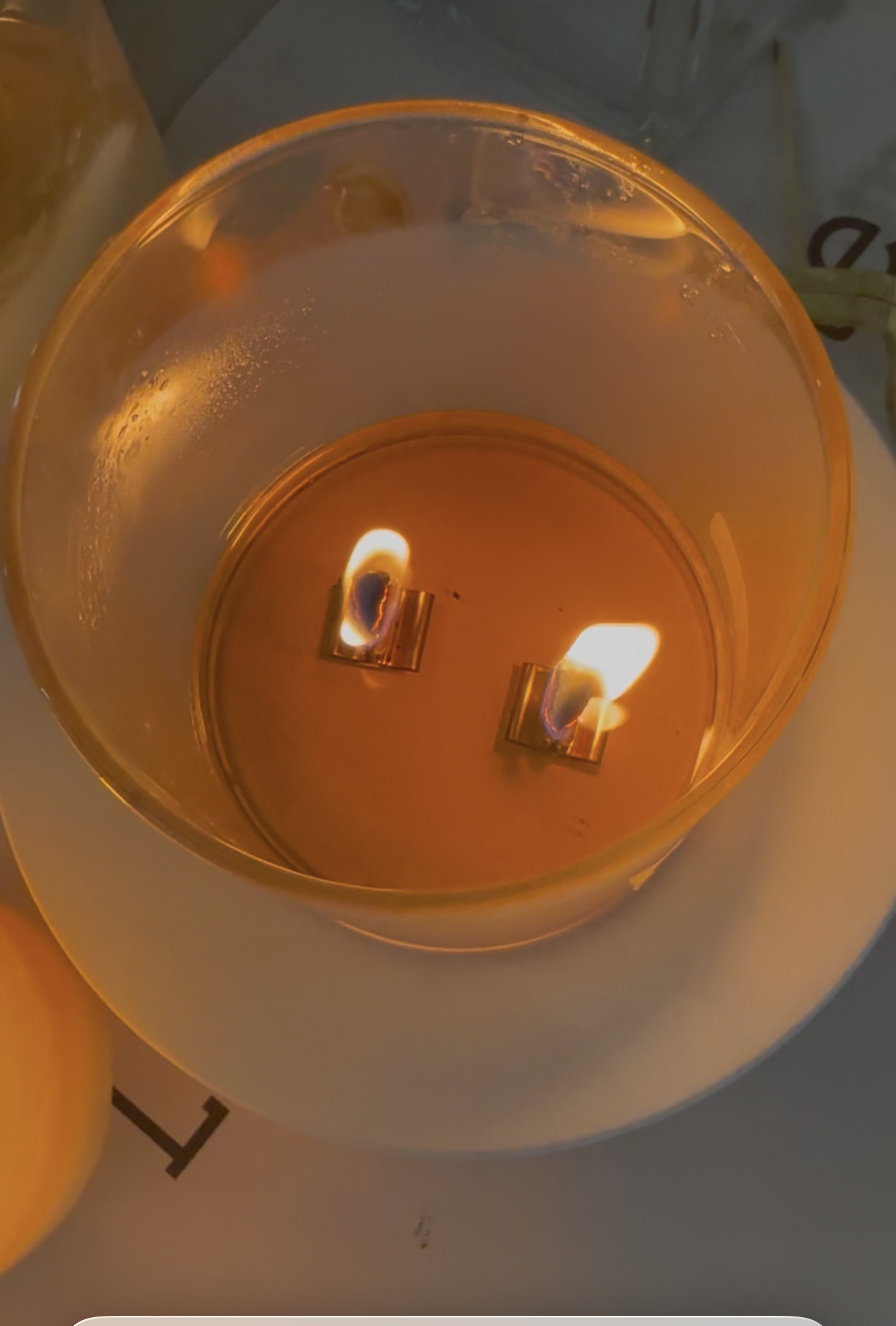 Two wooden wicks burning in a glass candle vessel with melted wax visible